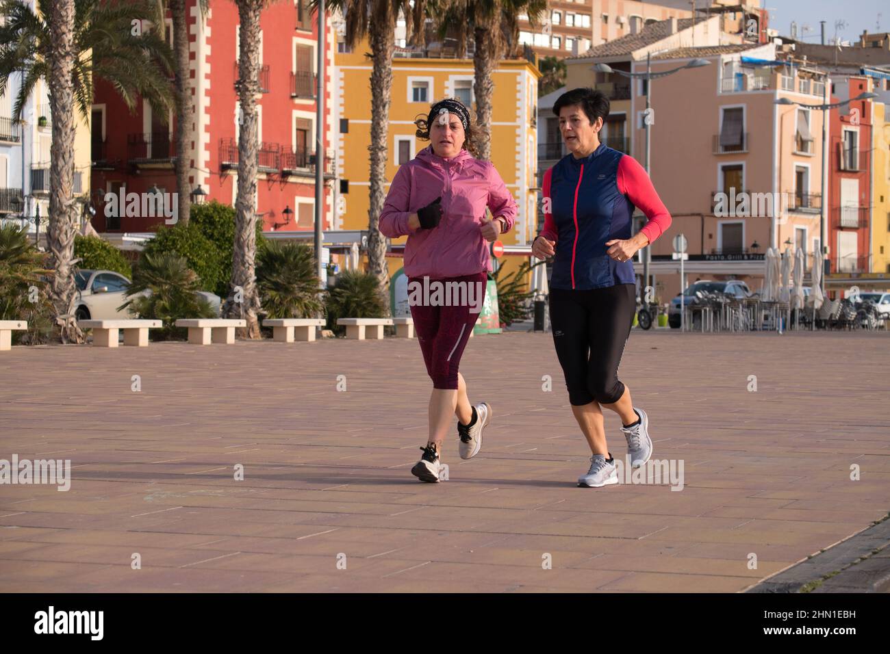 Villajoysta Alicante Spain 02.13.22 zwei ältere Damen tragen Leggings und langärmelige winddichte Westen beim Joggen entlang einer Fußgängerzone. Palmen. Stockfoto