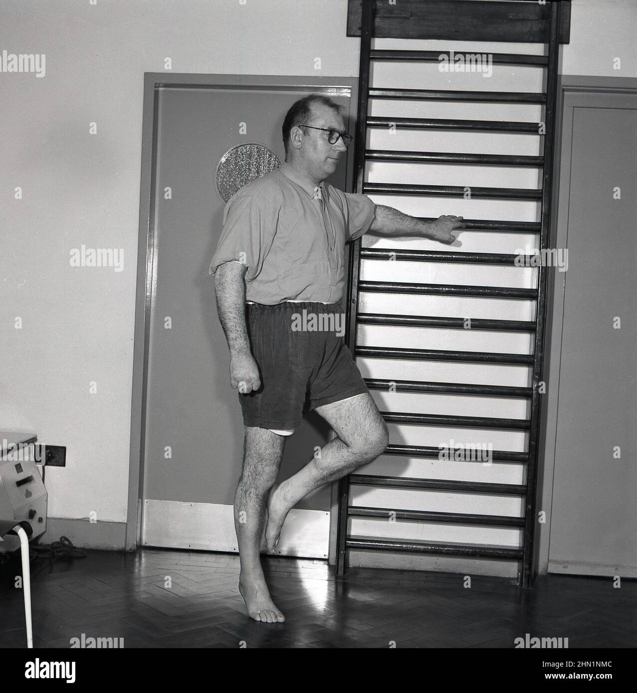 1950s, historisch, ein männlicher Arbeiter, der im Rahmen einer betrieblichen medizinischen oder gesundheitlichen Überprüfung Balancierübungen mit einer Hand auf einem Parrelel-Stab und einem Knie nach oben macht, Abbey Works, Port Talbot, Wales, Großbritannien. Stockfoto