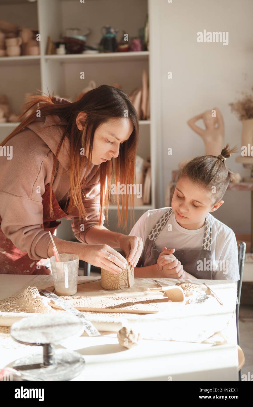 Vertikale Aufnahme einer weiblichen Keramikerin, die ihrer jungen Schülerin während des Töpferunterlasses hilft Stockfoto