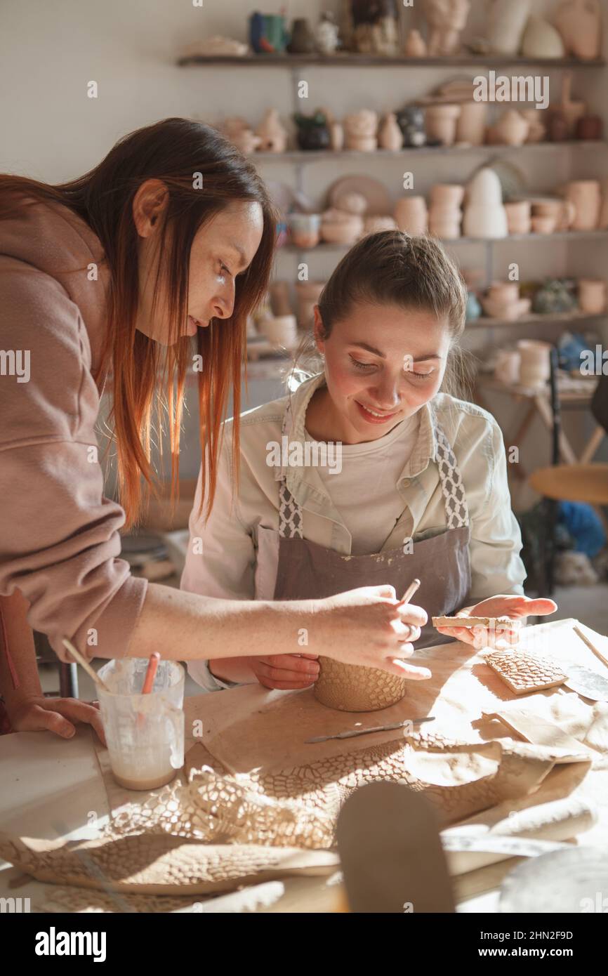 Vertikale Aufnahme einer professionellen Keramikerin, die ihrer Schülerin bei der Herstellung einer Tasse hilft Stockfoto