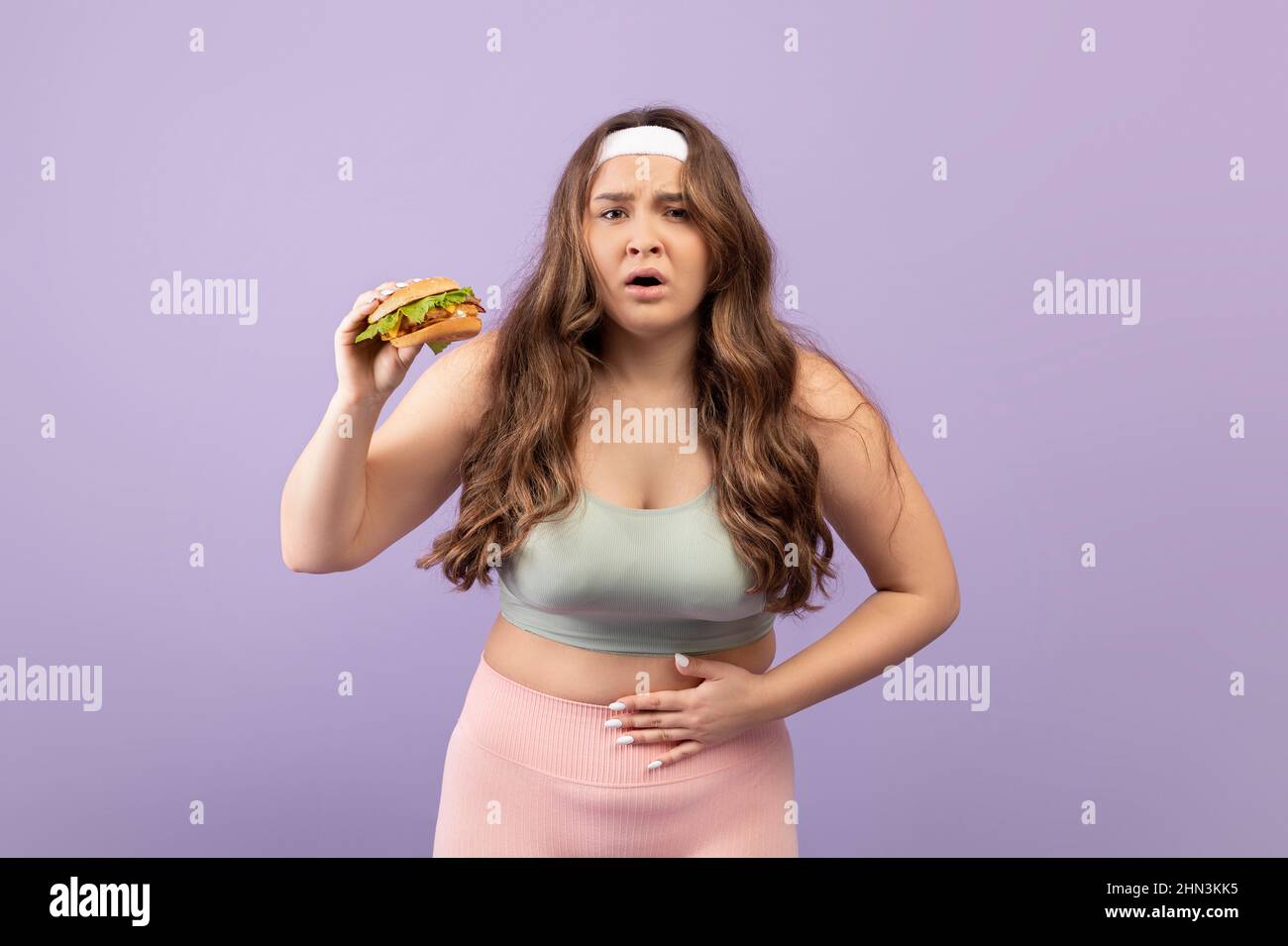 Unglückliche junge europäische plus size Dame hält Burger, drückt die Hand auf ihren Magen leiden unter Schmerzen Stockfoto