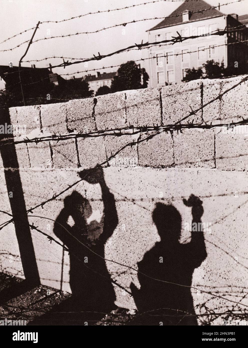 Vintage-Foto der Berliner Krise von 1961: Der Mauerschatten zweier West-Berliner, die mit Freunden über die Grenze winken, fällt symbolisch auf Con Stockfoto