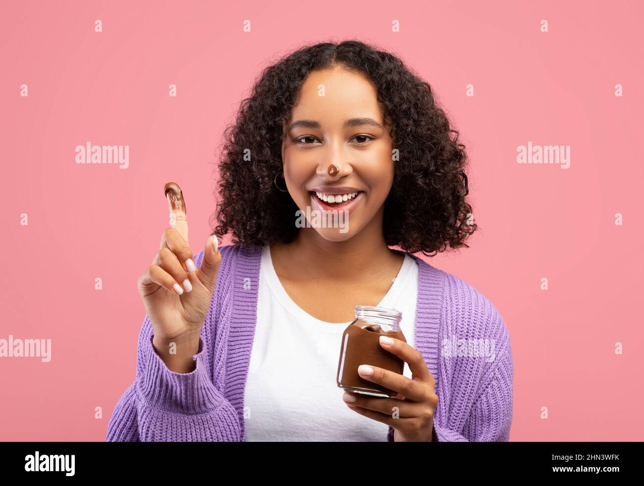 Lustige junge afroamerikanische Frau, die Schokoladenpaste aus dem Glas mit dem Finger isst, schmutzige Nase hat und Nachtisch genießt Stockfoto