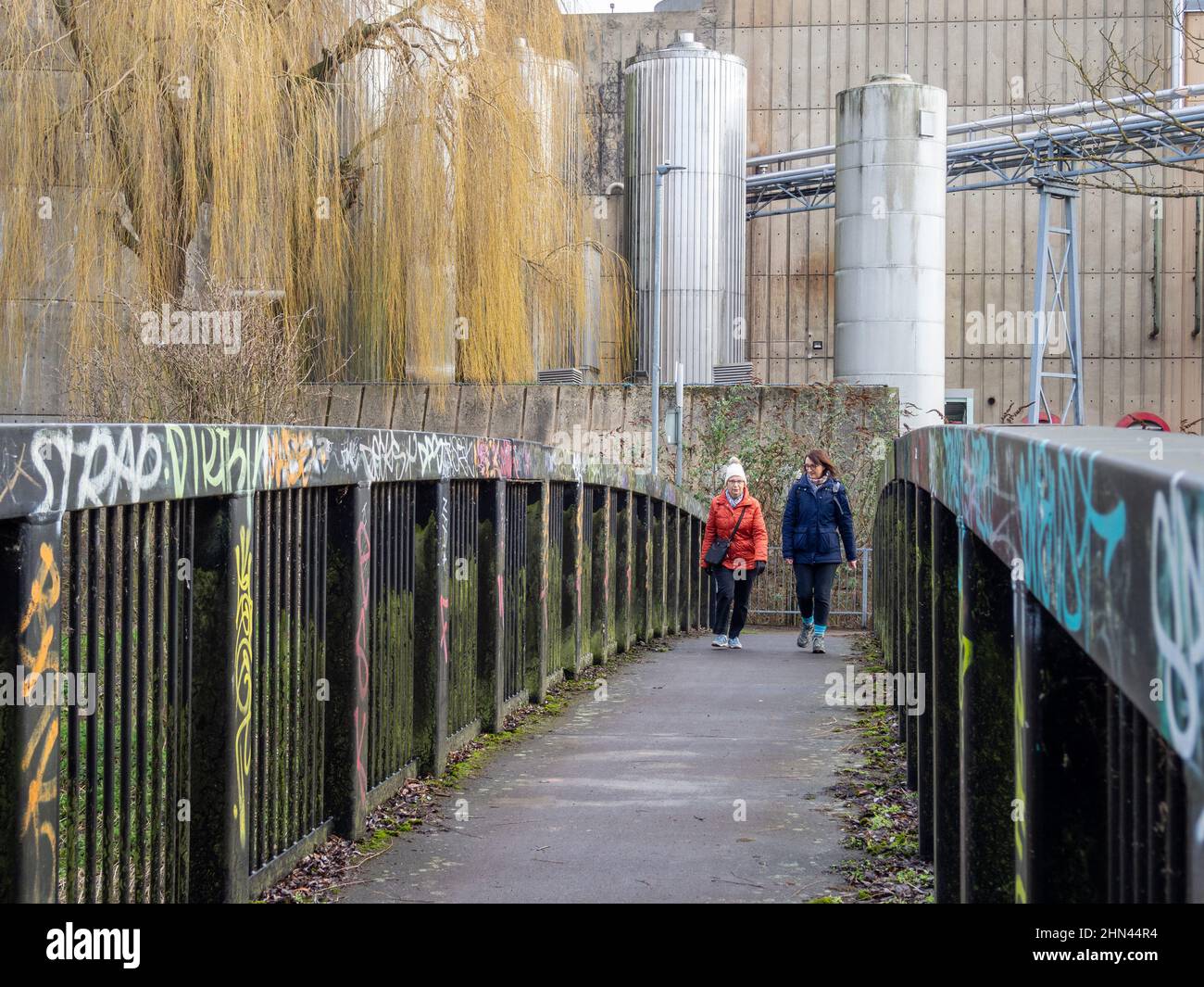 Ein Graffiti sprühte eine Fußgängerbrücke vor einem industriellen Hintergrund, Northampton, Großbritannien; zwei Frauen eilten über die Brücke Stockfoto