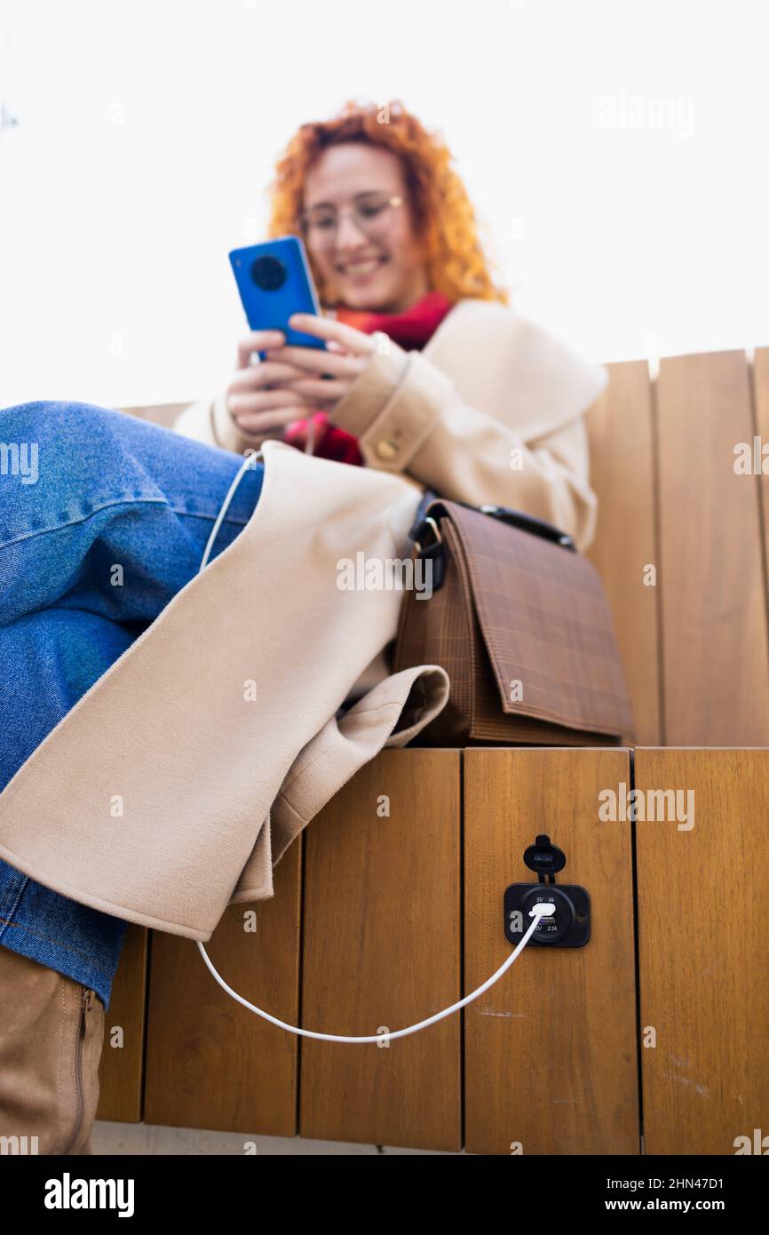 Junge Frau lädt ihr Smartphone auf einer intelligenten Bank, die sauberen Strom verwendet Stockfoto