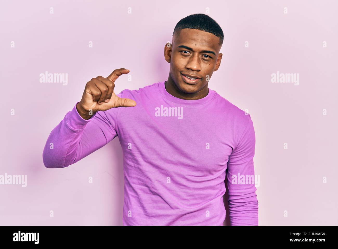 Junger schwarzer Mann trägt lässigen rosa Pullover lächelnd und zuversichtlich gestikulieren mit Hand tun kleine Größe Zeichen mit Fingern suchen und die Kamera. Measu Stockfoto