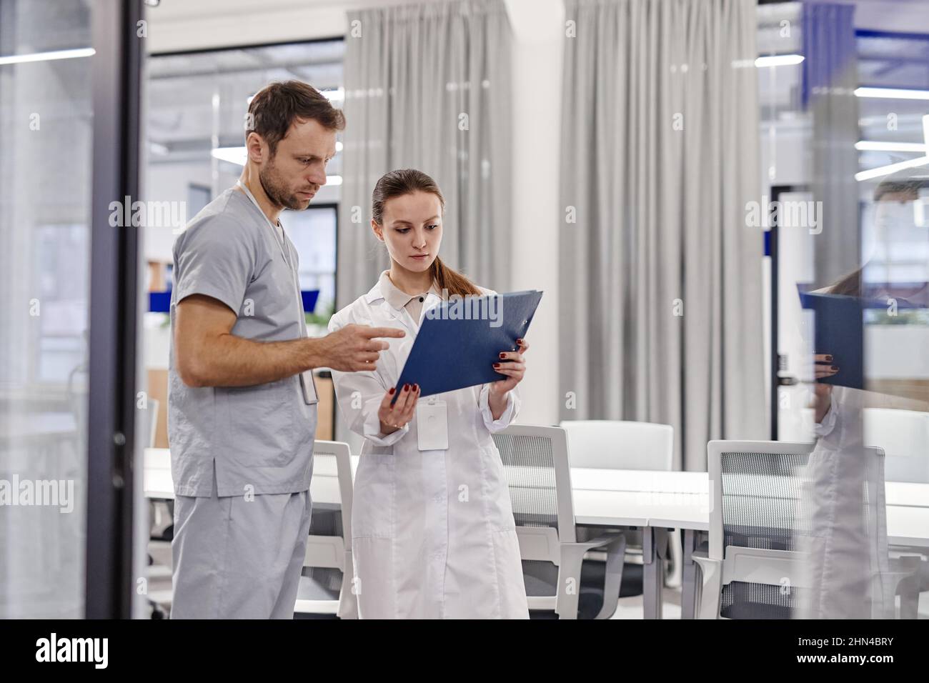 Junge Hausärztin im Labormantel, die einem Kollegen ein medizinisches Dokument vorzeigt, während sie sich mit ihm über Informationen berät Stockfoto