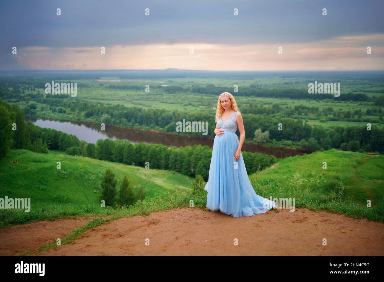 Schwangere in blauem Kleid, grünen Hügeln, Fluss und Himmel Stockfoto