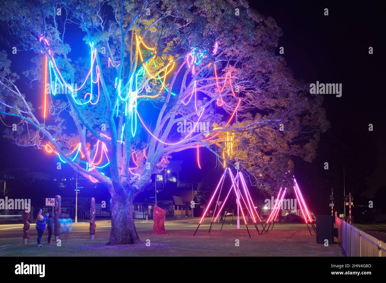 Farbenfrohe Lichtinstallationen in einem Park bei Nacht. Seilleuchten baumeln von einem Baum und dreieckige Lichtskulpturen stehen im Hintergrund Stockfoto