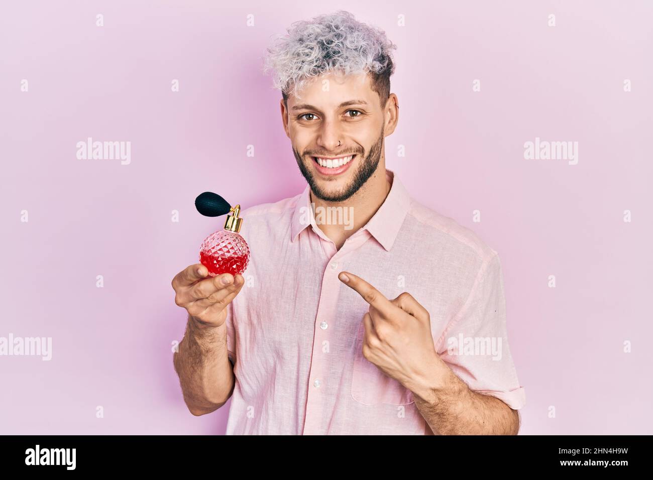 Junger hispanischer Mann mit modern gefärbtem Haar, das Luxusparfüm hält, lächelt glücklich und zeigt mit Hand und Finger Stockfoto