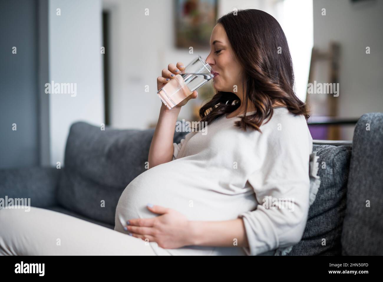 Schwangerin entspannt sich zu Hause. Sie sitzt auf dem Bett und trinkt Wasser. Stockfoto