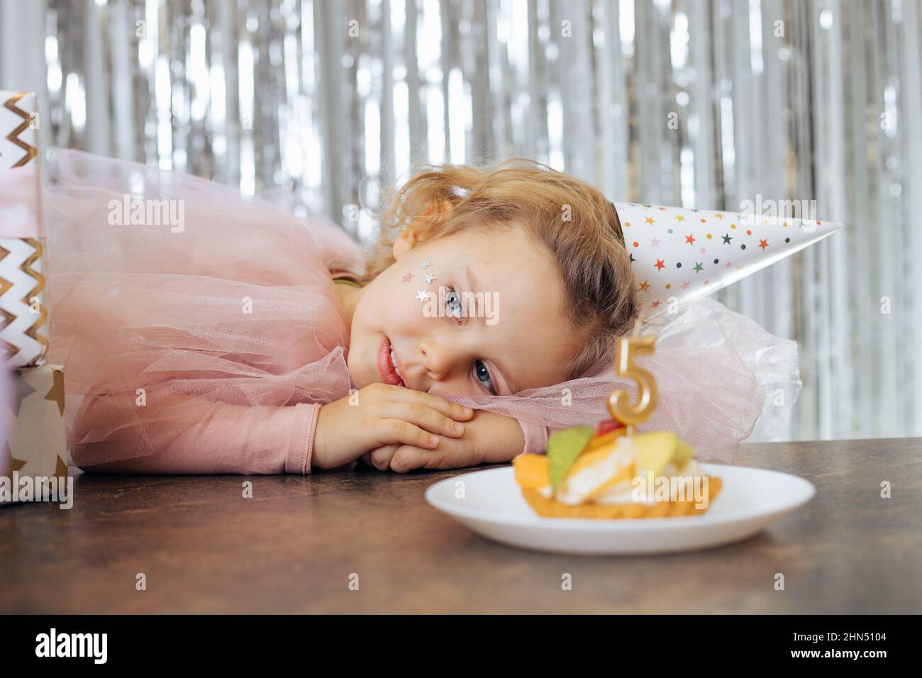 Kleines lächelndes Mädchen mit kurzen lockigen Haaren und Sternen im Gesicht in rosa poofy Kleid und Kappe legte den Kopf auf den Tisch faltende Hände. Stockfoto