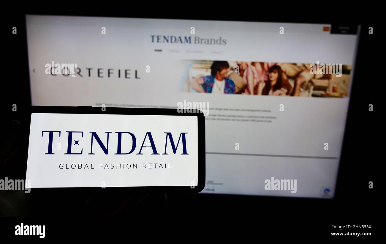 Person, die Mobiltelefon mit Logo des spanischen Modeunternehmens Tendam Brands S.A.U. auf dem Bildschirm vor der Business-Webseite hält. Konzentrieren Sie sich auf die Telefonanzeige. Stockfoto