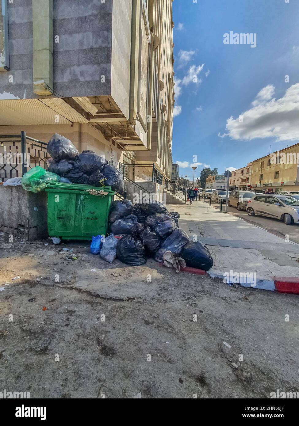 Volle Mülltonne in der Straße auf dem Bürgersteig in der Nähe des ElQods Zentrums in Cheraga, Algier. Menschen gehen und cas geparkt Stockfoto