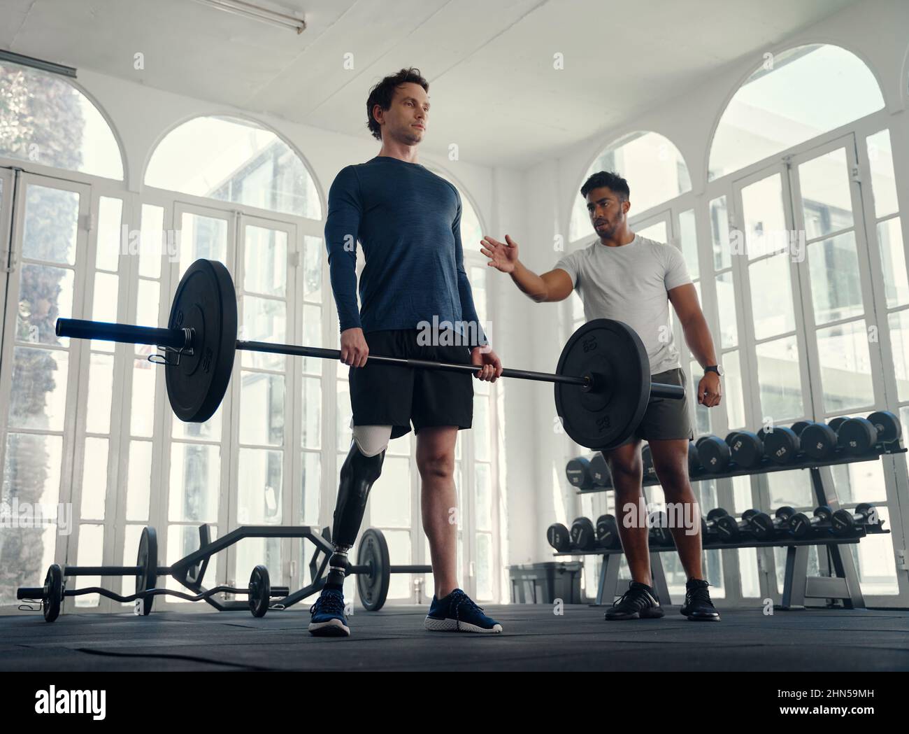Unterschiedlich trainierte Sportler Gewichtheben mit seinem Trainer in der Turnhalle. Mann mit Beinprothese, der von seinem Trainer trainiert wird Stockfoto