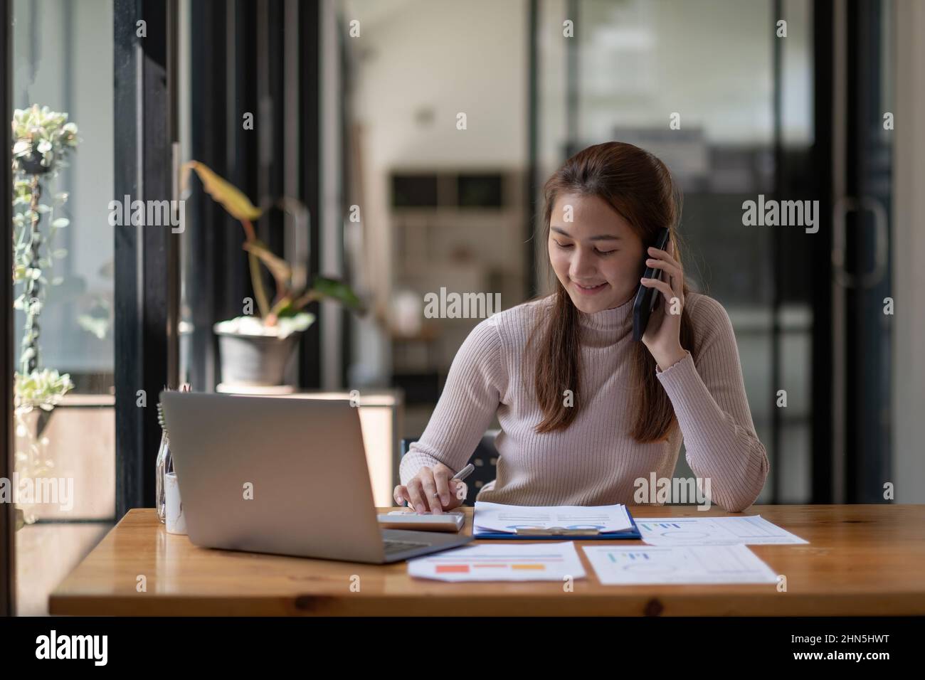 Porträt einer jungen asiatischen Frau, die an ihrem Schreibtisch sitzt und mit Taschenrechner und Papierkram arbeitet, während sie im modernen Büro mit dem Mobiltelefon telefoniert. Stockfoto