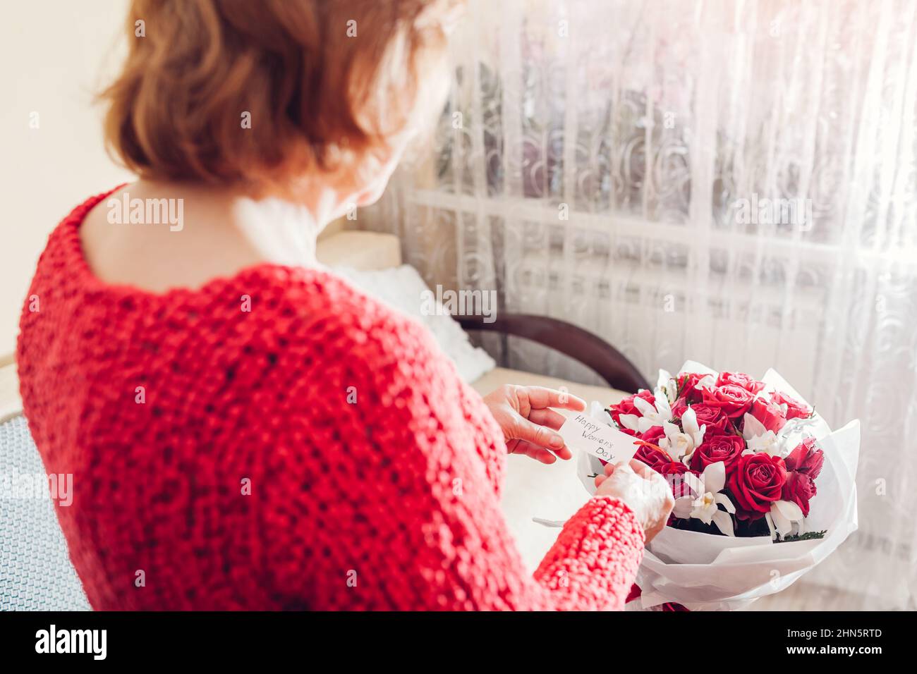 Happy Womens Day, 8 März Geschenk. Ältere Frau liest die Grußkarte im Strauß roter Rosen und Orchideen-Blumen zu Hause. Überraschung für den Urlaub Stockfoto