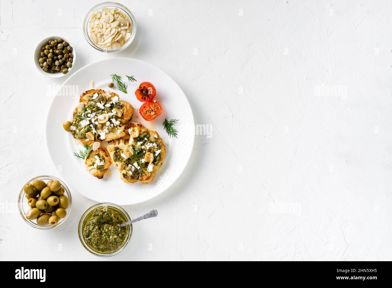 Blumenkohlesteak mit Gewürzen, Chimichurri-Sauce, Mandelflocken, Oliven, gebratenen Kirschtomaten und Kapern auf einem weißen Teller. Vegetarische Gerichte. Weiß, Bac Stockfoto