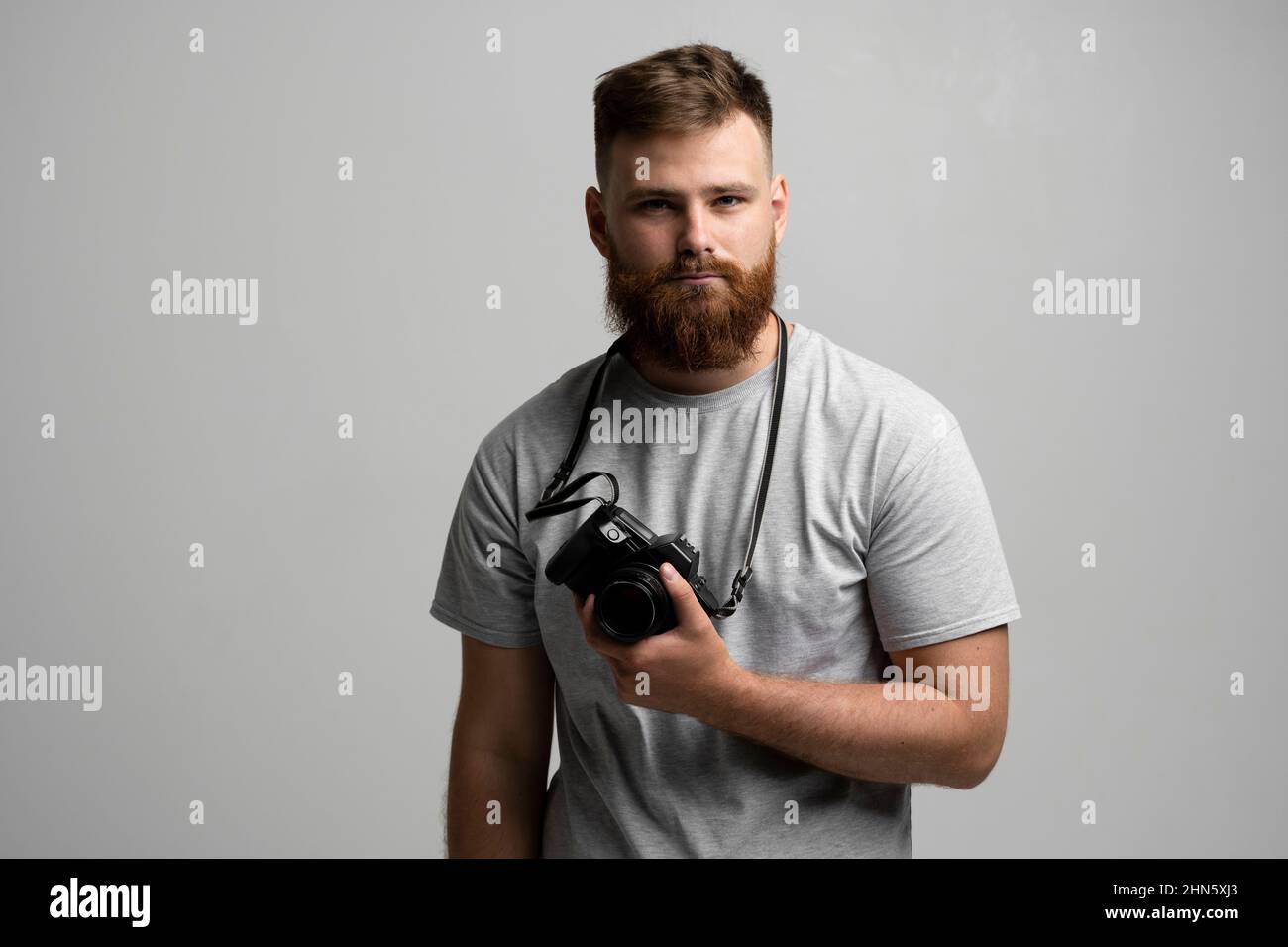 Porträt des bärtigen professionellen Fotografen mit dslr-Kamera schaut direkt in die Kamera isoliert auf grauem Hintergrund. Stockfoto