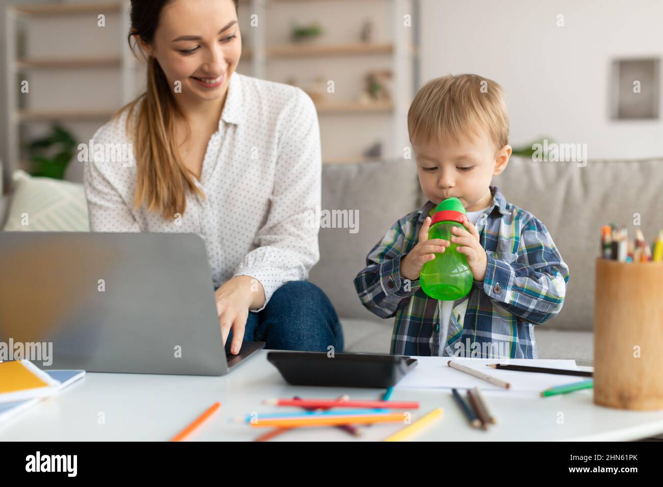 Glückliche tausendjährige Mutter, die am Laptop arbeitet, während ihr entzückender Sohn Wasser aus der Flasche trinkt und in der Nähe der Mutter steht Stockfoto