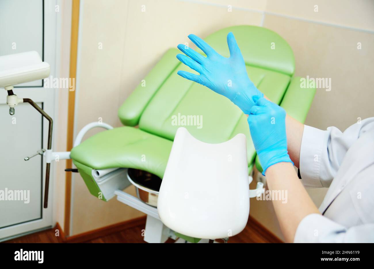 Der Gynäkologe, der Gummihandschuhe anzieht, bereitet sich auf die Untersuchung vor dem Hintergrund des gynäkologischen Stuhls vor. Stockfoto