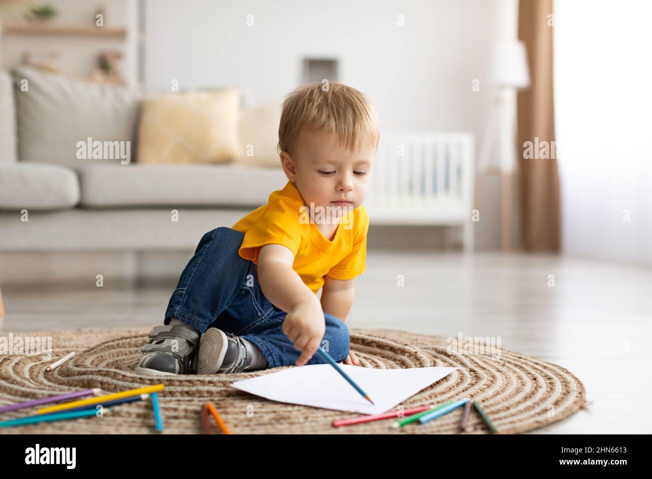 Kaukasischer kleiner Junge, der zu Hause ein Bild mit bunten Bleistiften zeichnet und auf dem Teppich im Wohnzimmer sitzt Stockfoto