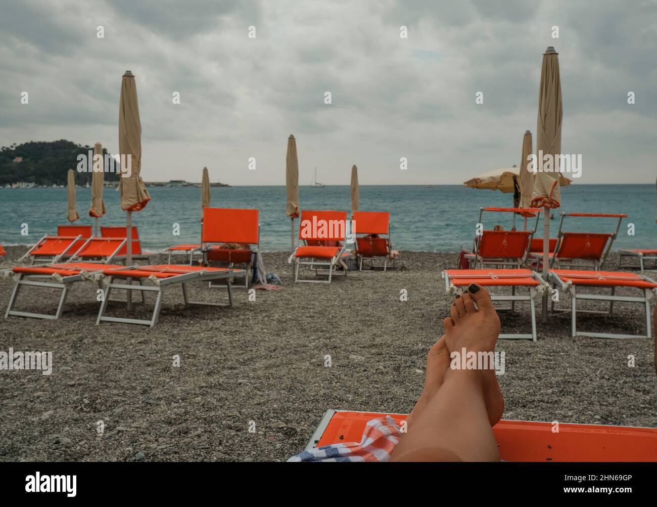 Orange Liegestühle und Sonnenschirme am Strand über Meer und Horizont. Die Beine der Frau auf dem Strandstuhl. Sommerurlaub. Entspannen Sie sich am Strand Stockfoto