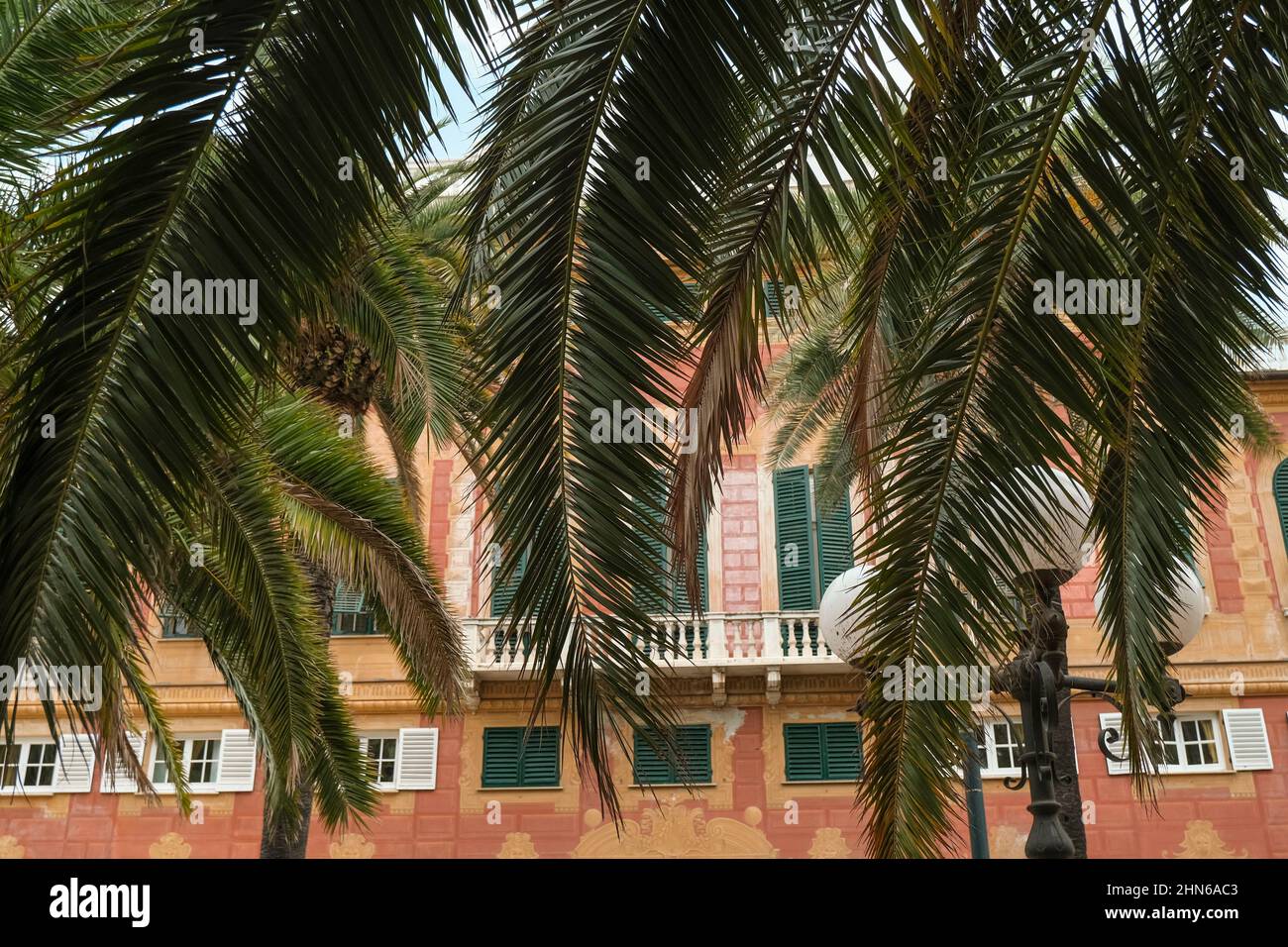 Palmenblätter und Äste gegenüber dem orangefarbenen und roten Hotelgebäude mit grünen Fensterläden am Himmel. Sommerurlaub Stimmung, Hintergrund. Mittelmeer Stockfoto