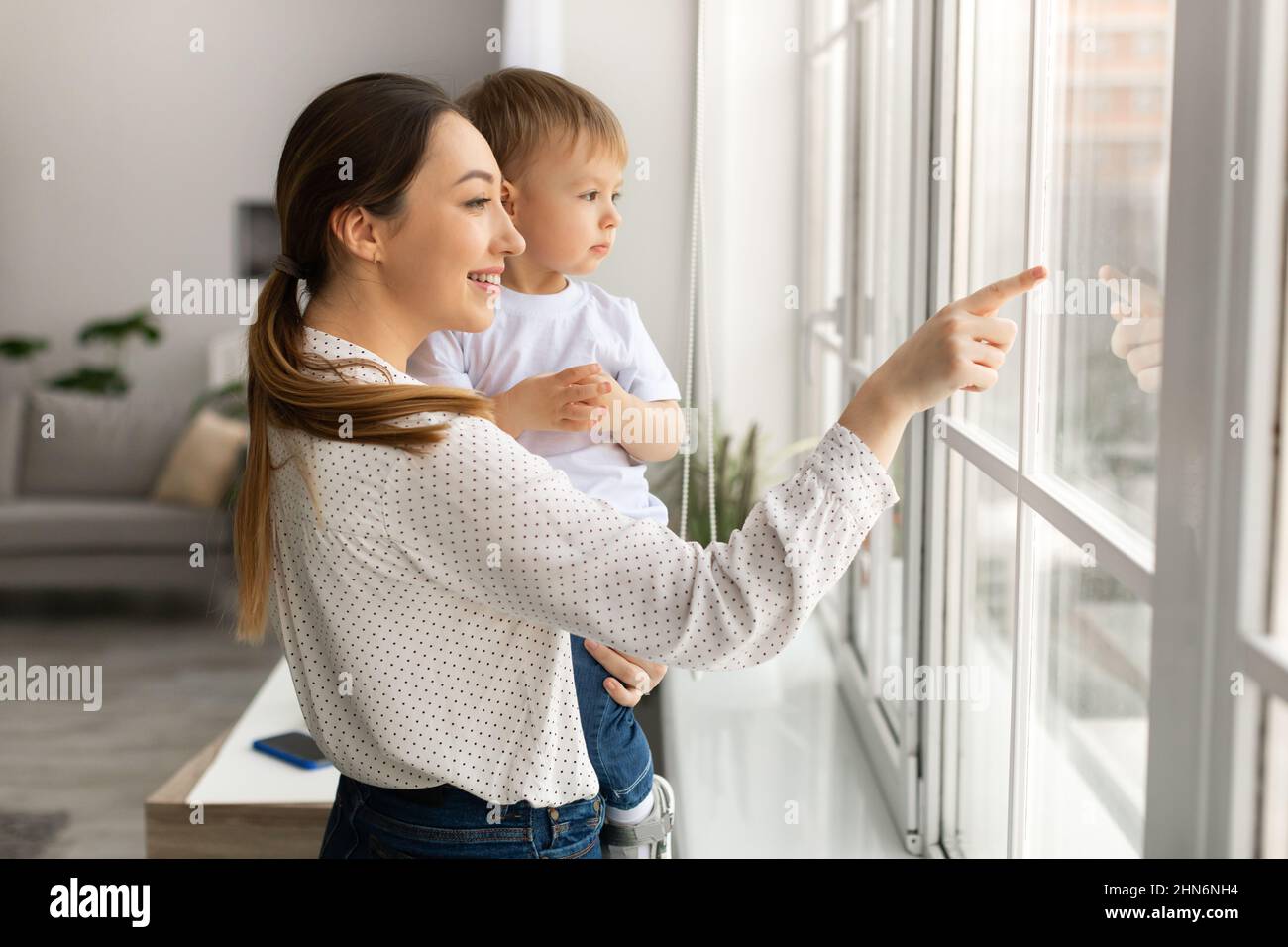 Glückliche junge Mutter hält den kleinen Sohn an den Händen, steht am Fenster und zeigt ihm die Welt draußen Stockfoto