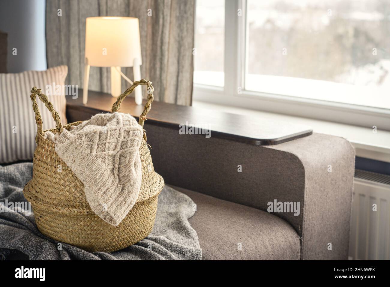 Strohkorbkorb mit Strick auf Sofa im Innenraum. Bambus Korb stilvolle Innenausstattung Öko-Design handgefertigt. Einrichtung wie zu Hause. Stockfoto