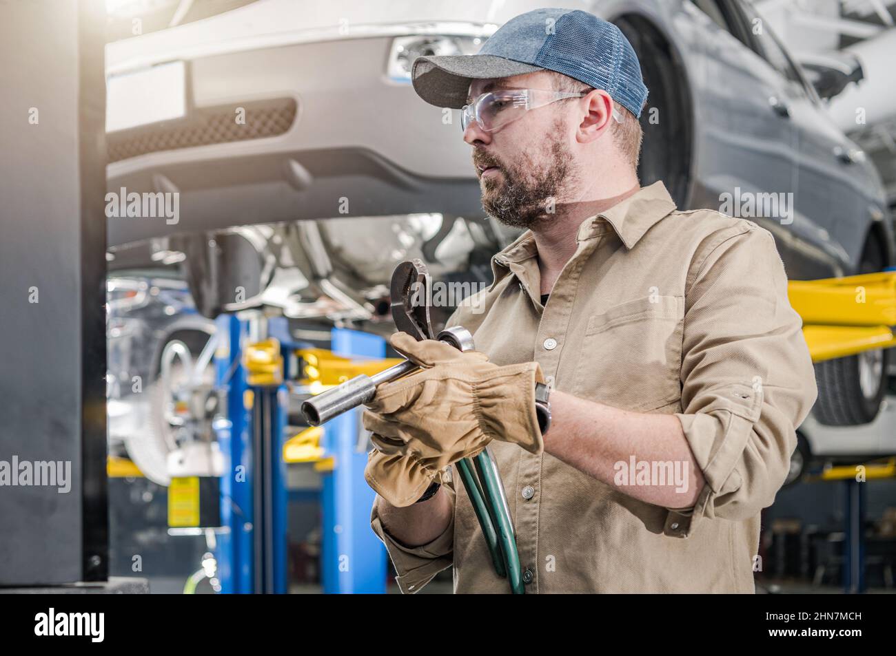 Kaukasische professionelle Kfz-Mechaniker Auswahl der richtigen Werkzeuge für einen Job. Kaputte Autos auf einem Automobil heben sich direkt hinter ihm. Transport Th Stockfoto