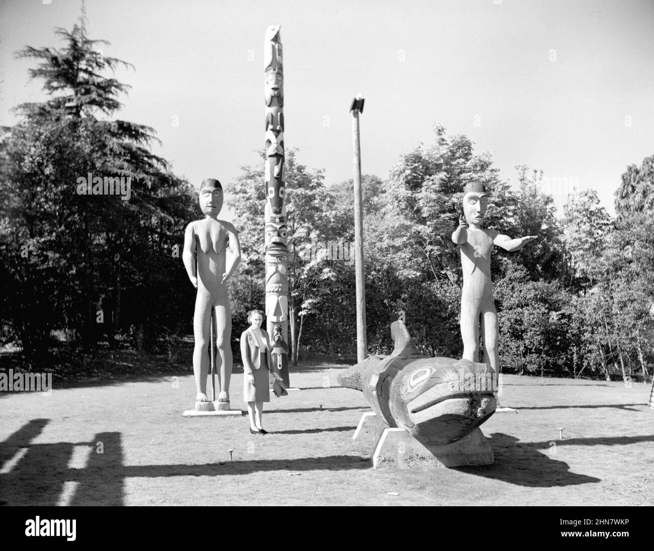 Vintage Schwarz-Weiß-Fotografie einer Frau, die neben einem Totempfähle der Nordwestküste und Skulpturen im Thunderbird Park Ca. 1940 in Victoria, British Columbia, Kanada Stockfoto