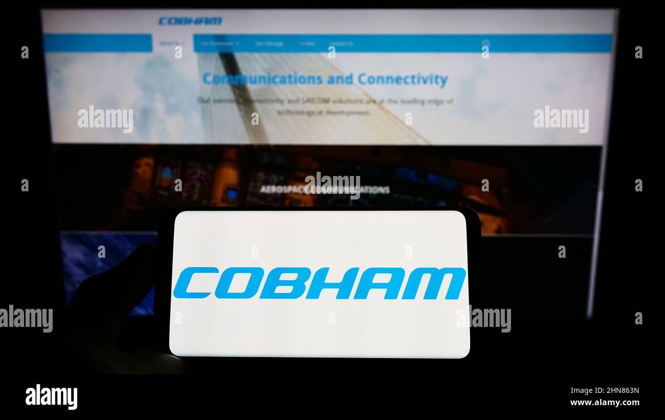 Person, die Mobiltelefon mit dem Logo des britischen Luftfahrtunternehmens Cobham Ltd auf dem Bildschirm vor der Business-Webseite hält. Konzentrieren Sie sich auf die Telefonanzeige. Stockfoto