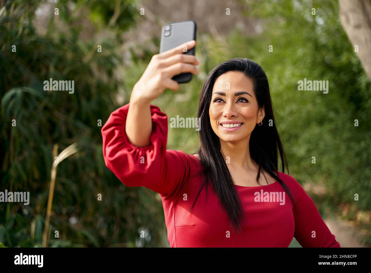 Glückliche hispanische Frau in roten Kleidern, die auf dem Smartphone Selbstporträt macht, während sie auf der Straße mit grünen Pflanzen in der Stadt steht Stockfoto