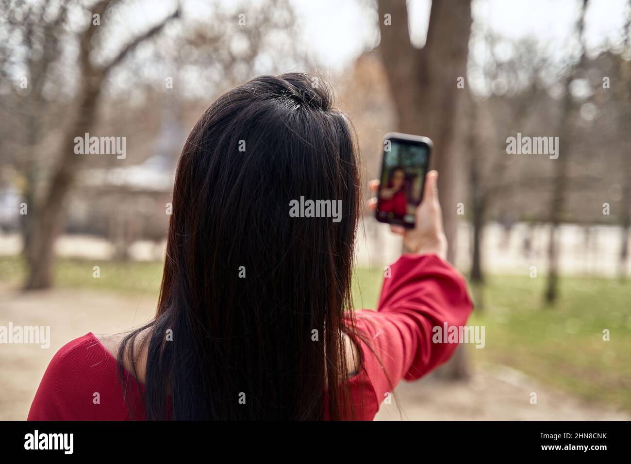 Rückansicht einer anonymen Frau mit schwarzen Haaren, die auf dem Smartphone ein Selbstporträt aufnahm, während sie im Park mit hohen Bäumen stand Stockfoto