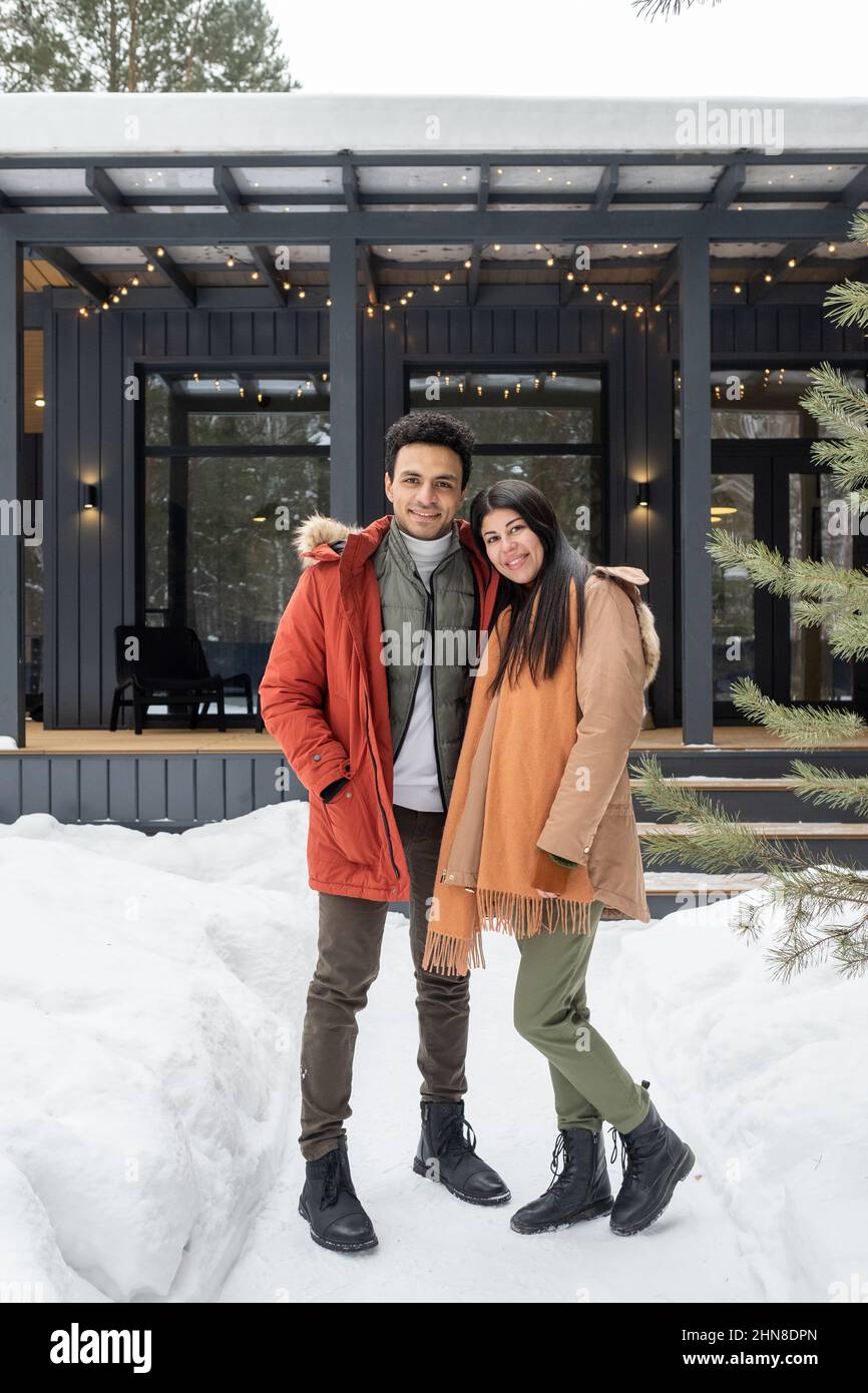 Porträt eines jungen Paares, das die Kamera umarmt und lächelt, während es während des Wintertages im Freien mit einem Haus im Hintergrund steht Stockfoto