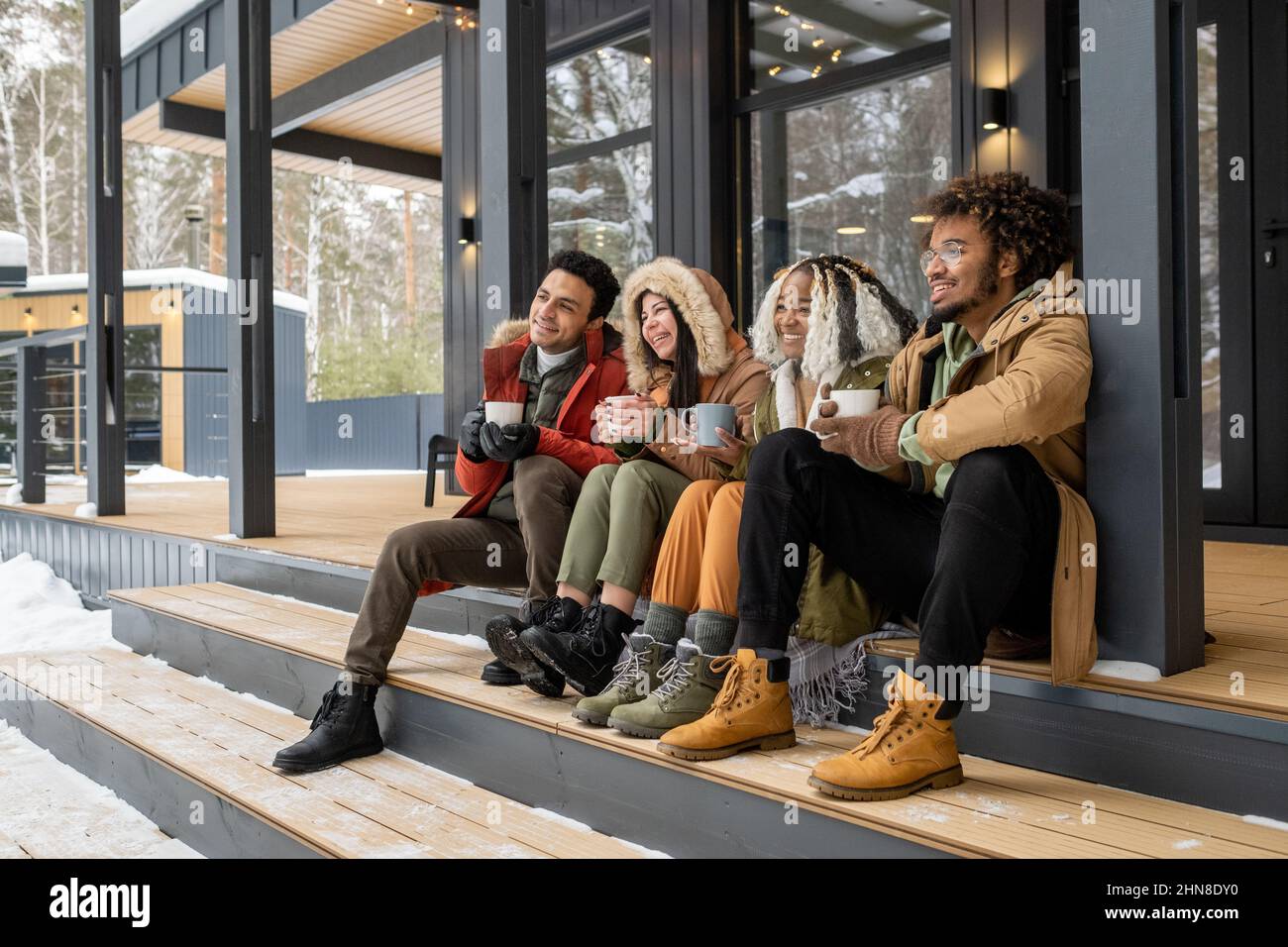 Eine Gruppe junger Freunde, die auf einer Treppe vor dem Landhaus sitzen, Kaffee trinken und den Winter genießen Stockfoto