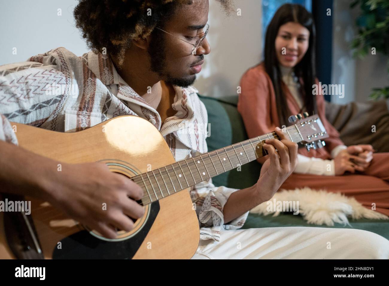 Afrikanischer junger Mann in einer Brille, der Gitarre spielt und ein Lied singt, während er auf dem Sofa sitzt, mit einer Frau im Hintergrund, die ihm zuhört Stockfoto