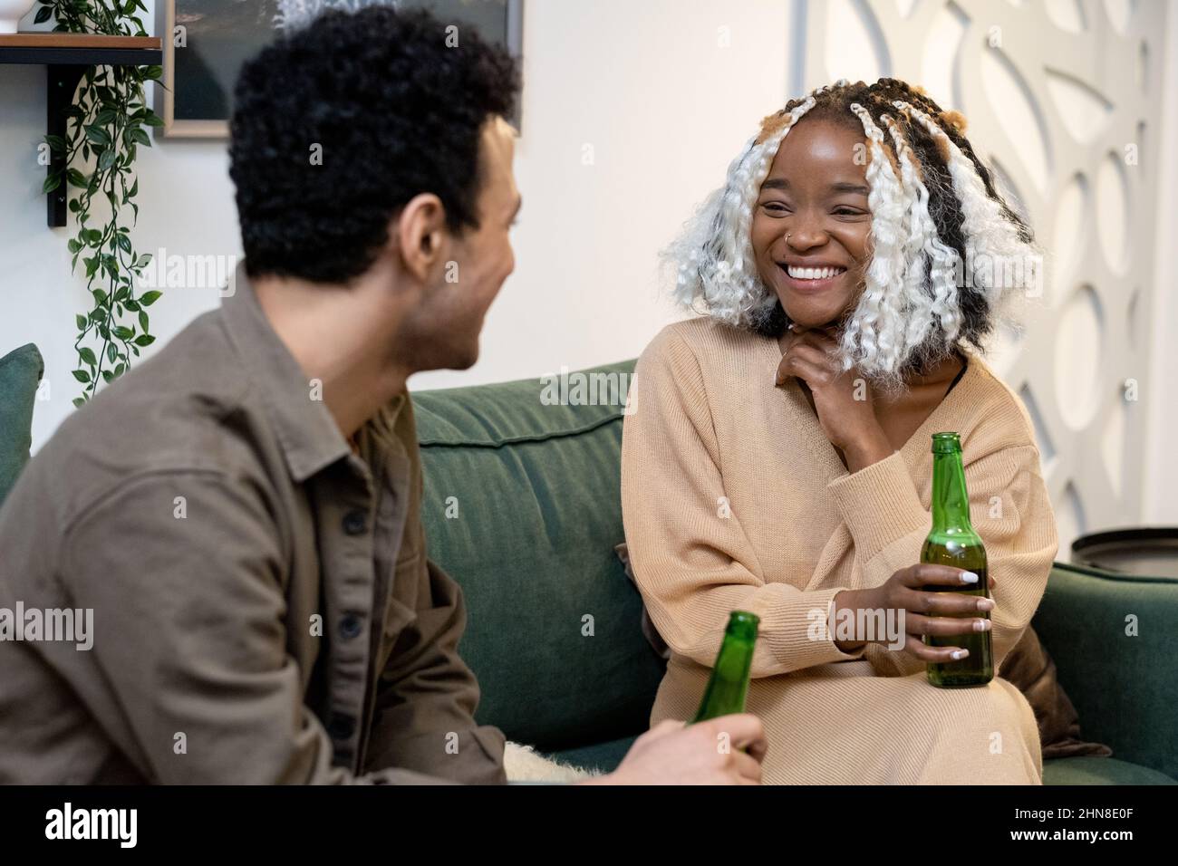 Ein multiethnisches junges Paar trinkt Bier und lacht, während es sich auf dem Sofa im Zimmer ausruht Stockfoto