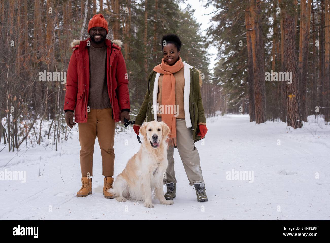 Porträt eines glücklichen afrikanischen Paares, das lächelt und die Kamera im Winterwald mit ihrem niedlichen Hund anschaut Stockfoto