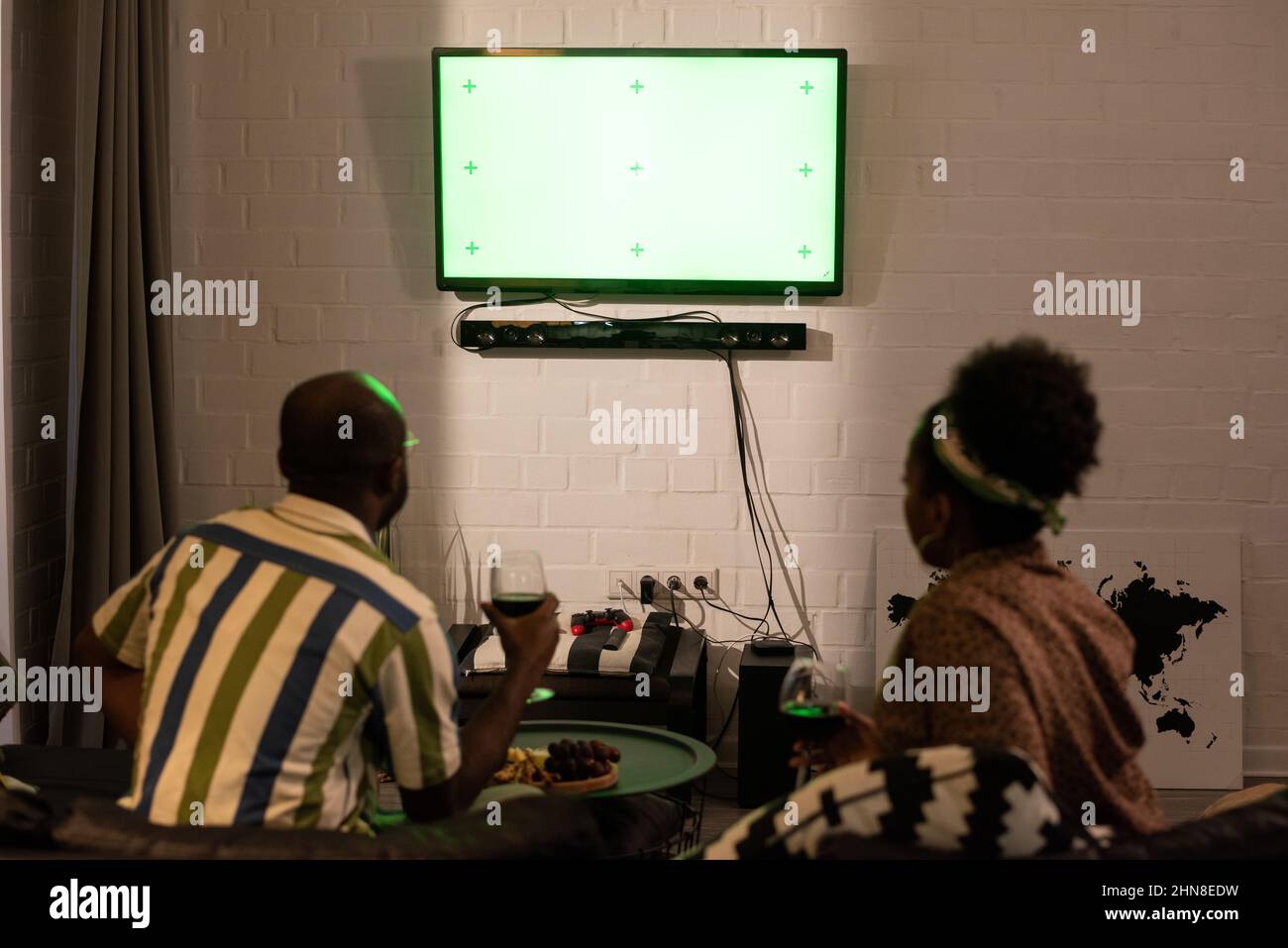 Rückansicht des afrikanischen Paares, das mit einem Glas Rotwein auf dem Sofa saß und auf den an der Wand hängenden Breitbildfernseher schaute, und am Abend einen Film sah Stockfoto