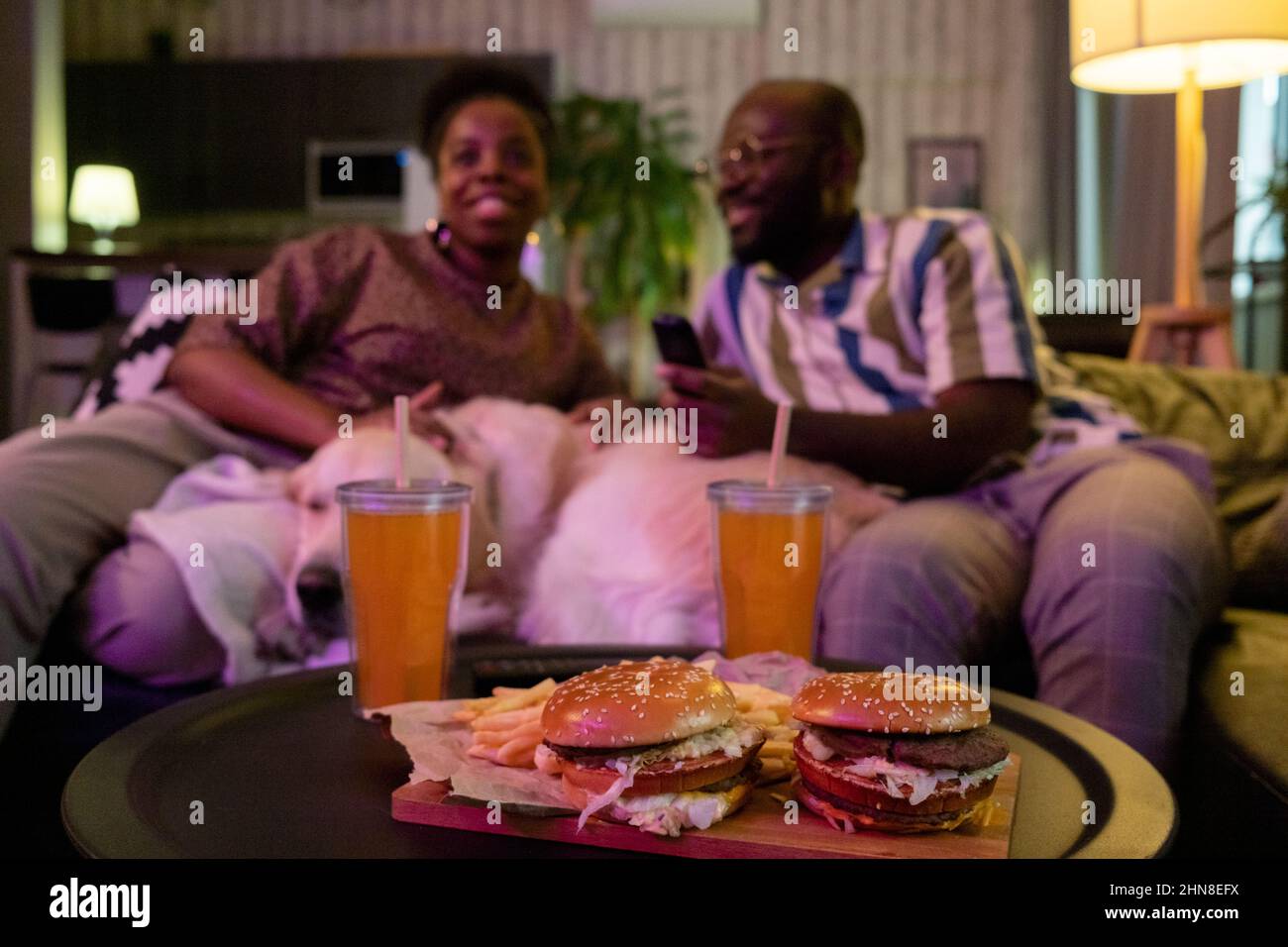 Nahaufnahme von Burgern und Limonade auf dem Tisch, während ein afrikanisches Paar auf dem Sofa im Hintergrund gemeinsam Fernsehen schaut Stockfoto