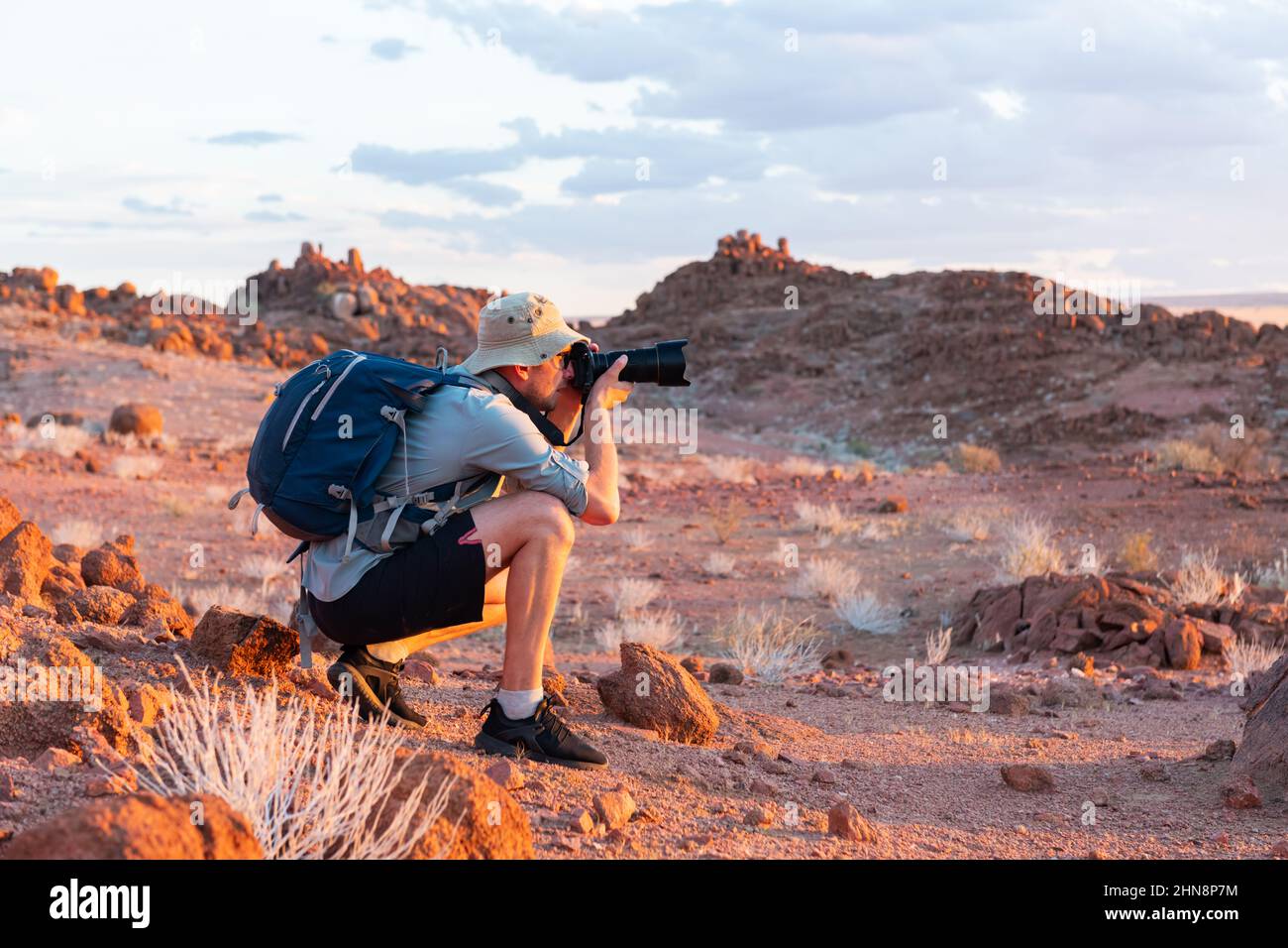 Fotograf, der in Felsen der Namib-Wüste, Namibia, Afrika fotografiert. Rote Berge und Sonnenuntergang Himmel im Hintergrund. Landschaftsfotografie Stockfoto
