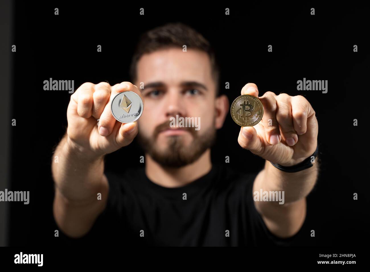 Junger bärtiger Mann mit virtueller Währung ethereum-Münze in einer Hand und Bitcoin-Münze in einer anderen auf schwarzem Hintergrund. Kryptowährung. Stockfoto