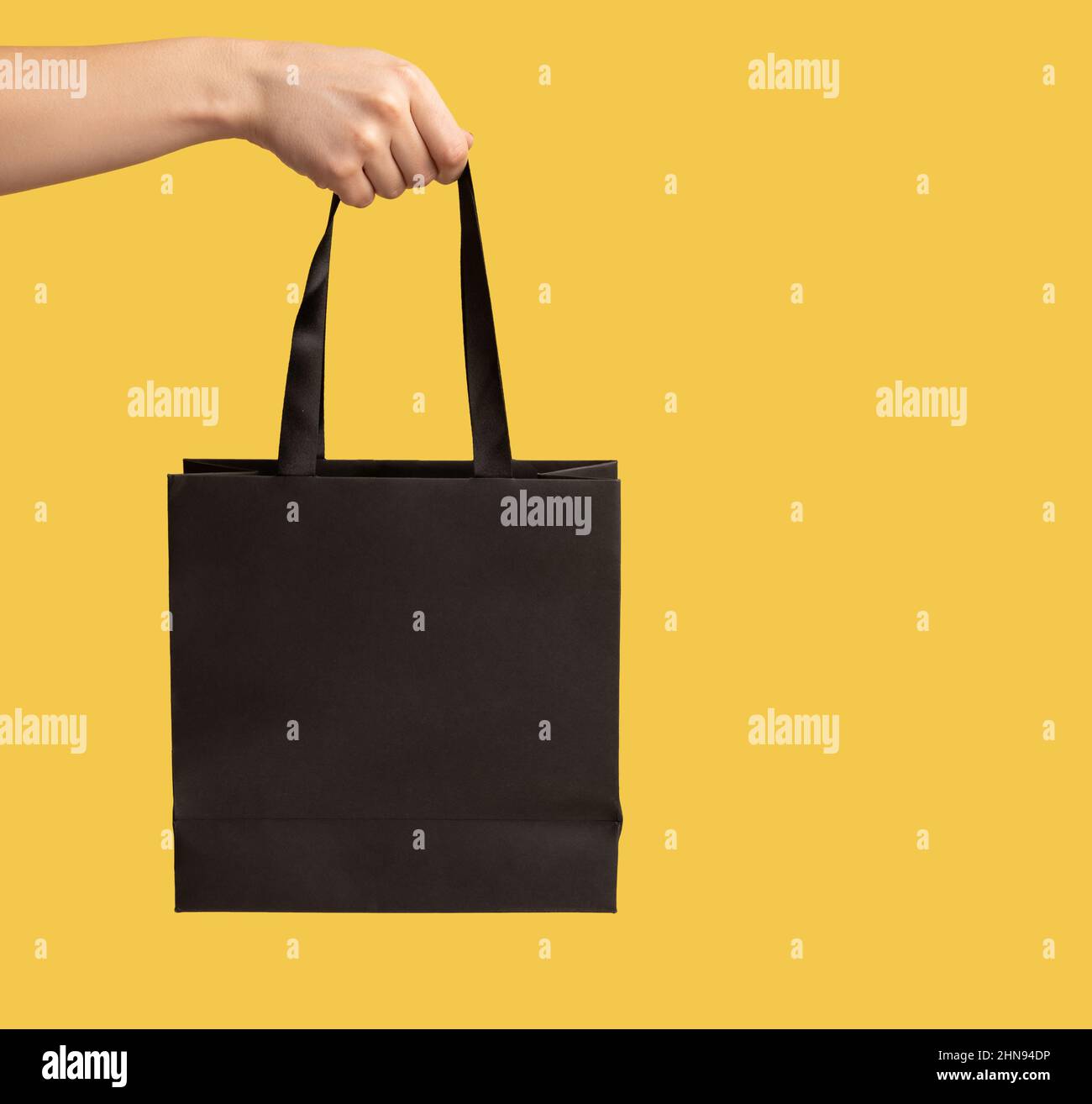 Nahaufnahme der Einkaufstasche im Frauenarm, Werbung für Käufe im Modegeschäft, weibliche Hand mit schwarzem Paket, kommerzielles Konzept. Innenaufnahme des Studios isoliert auf gelbem Hintergrund. Stockfoto