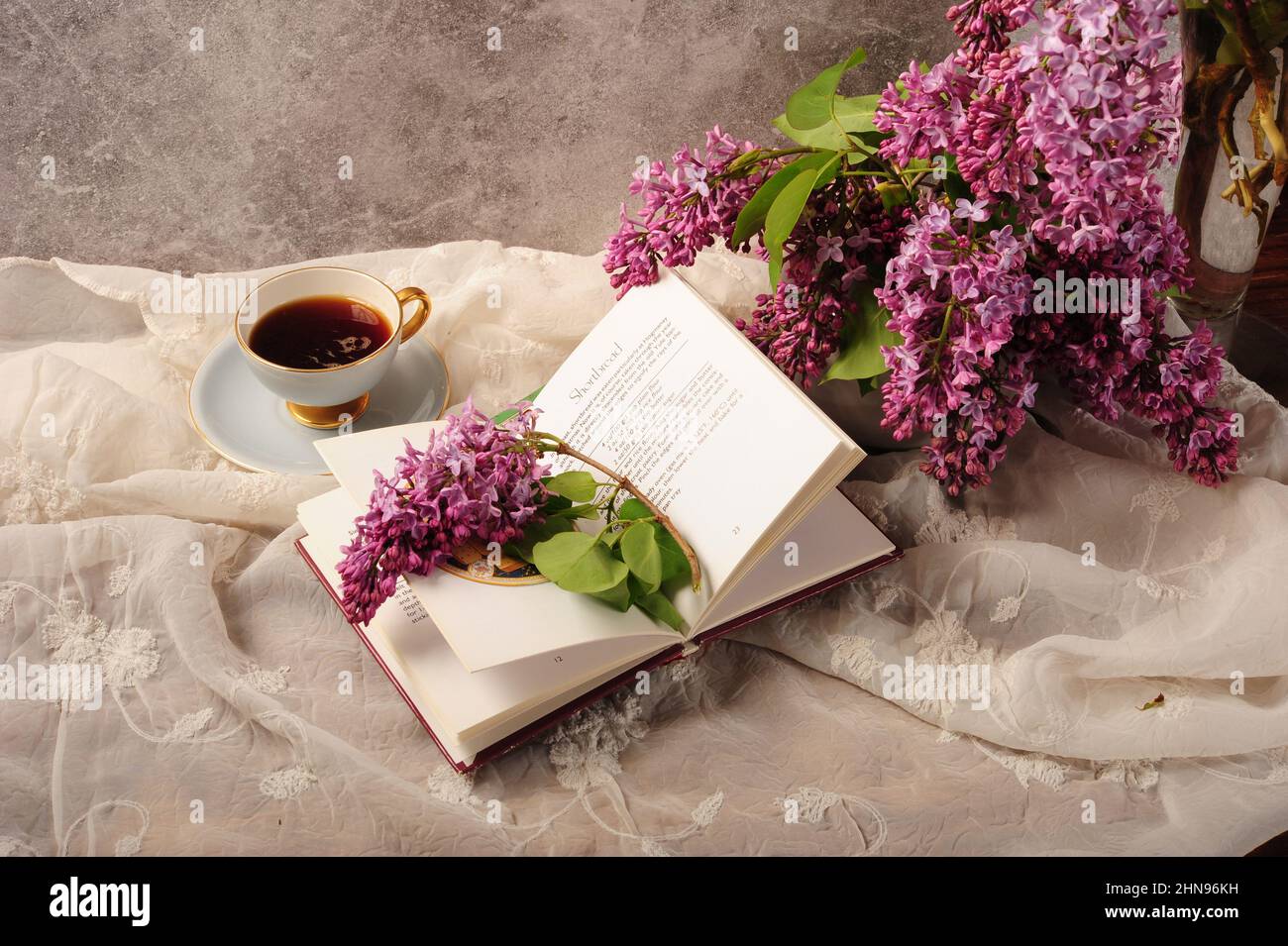 Kochbuch, Flieder und Kaffee auf besticktem weißen Tischtuch öffnen. Horizontales Foto mit Kopierbereich. Stockfoto