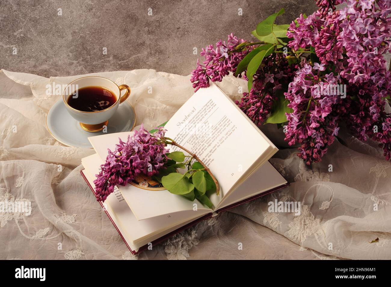 Kochbuch, Kaffee und Flieder-Bouquet wunderschön auf besticktem weißen Tischtuch arrangiert. Stockfoto