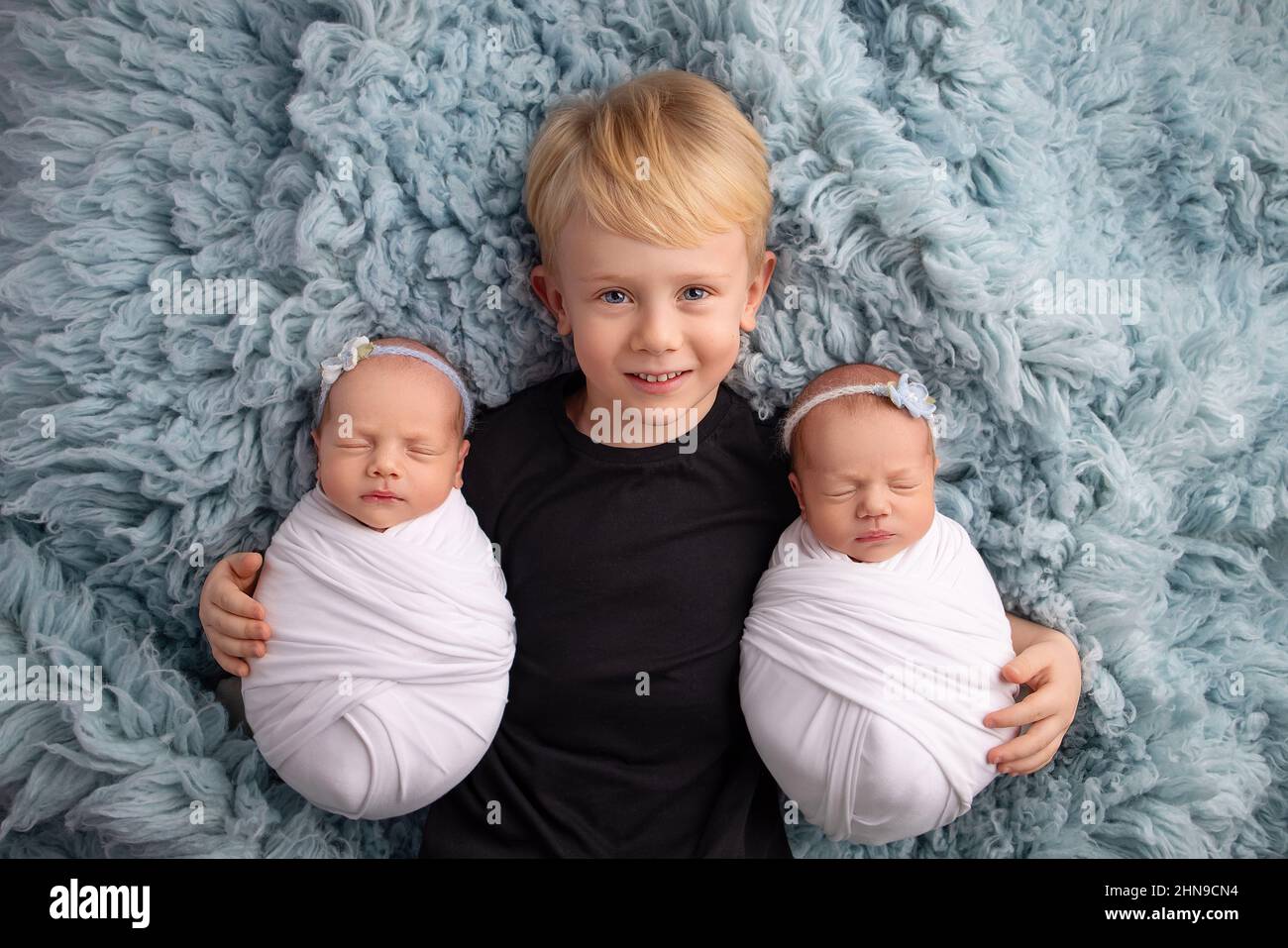 Kleine neugeborene Zwillingstündinnen in weißen Kokons auf blauem Hintergrund. Der ältere Bruder liegt zwischen ihnen und umarmt sanft seine Zwillingsschwestern. Stockfoto