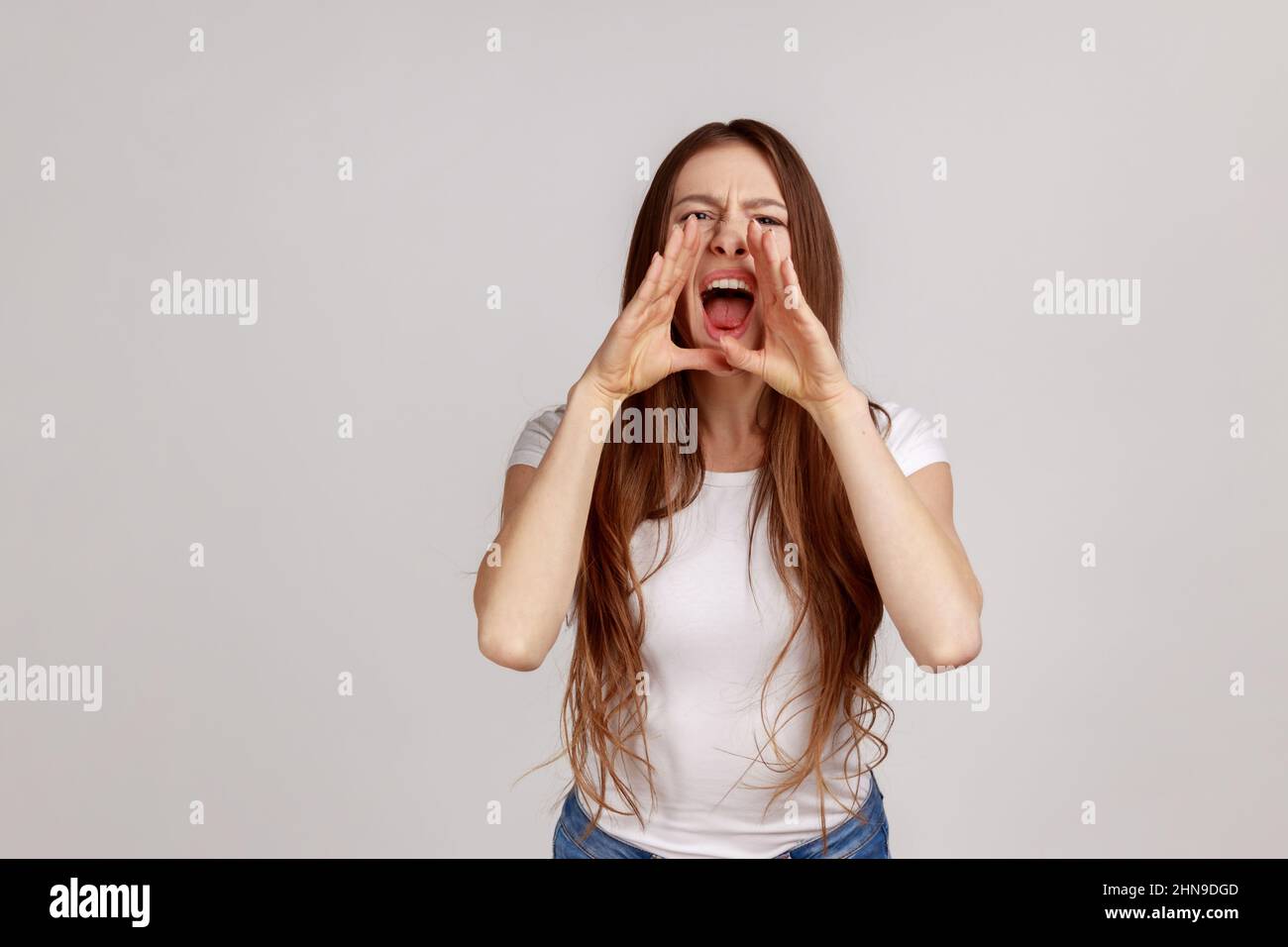 Porträt einer wütenden Frau mit dunklen Haaren, die steht und über schlechte Nachrichten oder Probleme schreit, ihre Hände in der Nähe des Mundes zu halten, mit weißem T-Shirt. Innenaufnahme des Studios isoliert auf grauem Hintergrund. Stockfoto