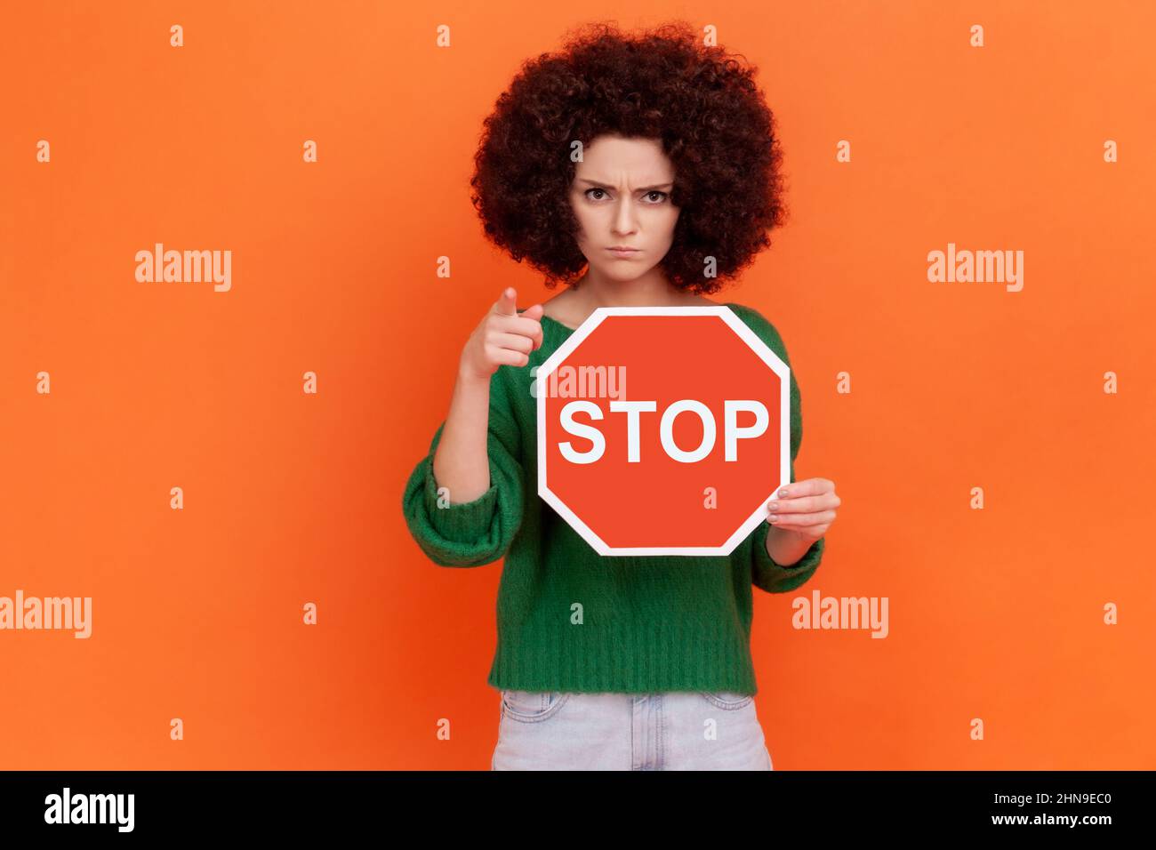 Hey, hör auf. Herrische Frau mit Afro-Frisur, die einen grünen Pullover im Casual-Stil trägt, der mit dem Finger auf die Kamera zeigt und in den Händen ein rotes Verkehrsstopschild zeigt. Innenaufnahme des Studios isoliert auf orangefarbenem Hintergrund. Stockfoto